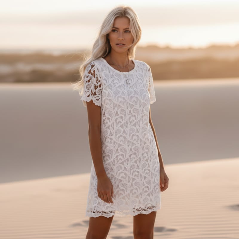White - A woman wearing a white short-sleeved lace shift dress with scalloped hems, standing in a desert sand dune landscape during golden hour.