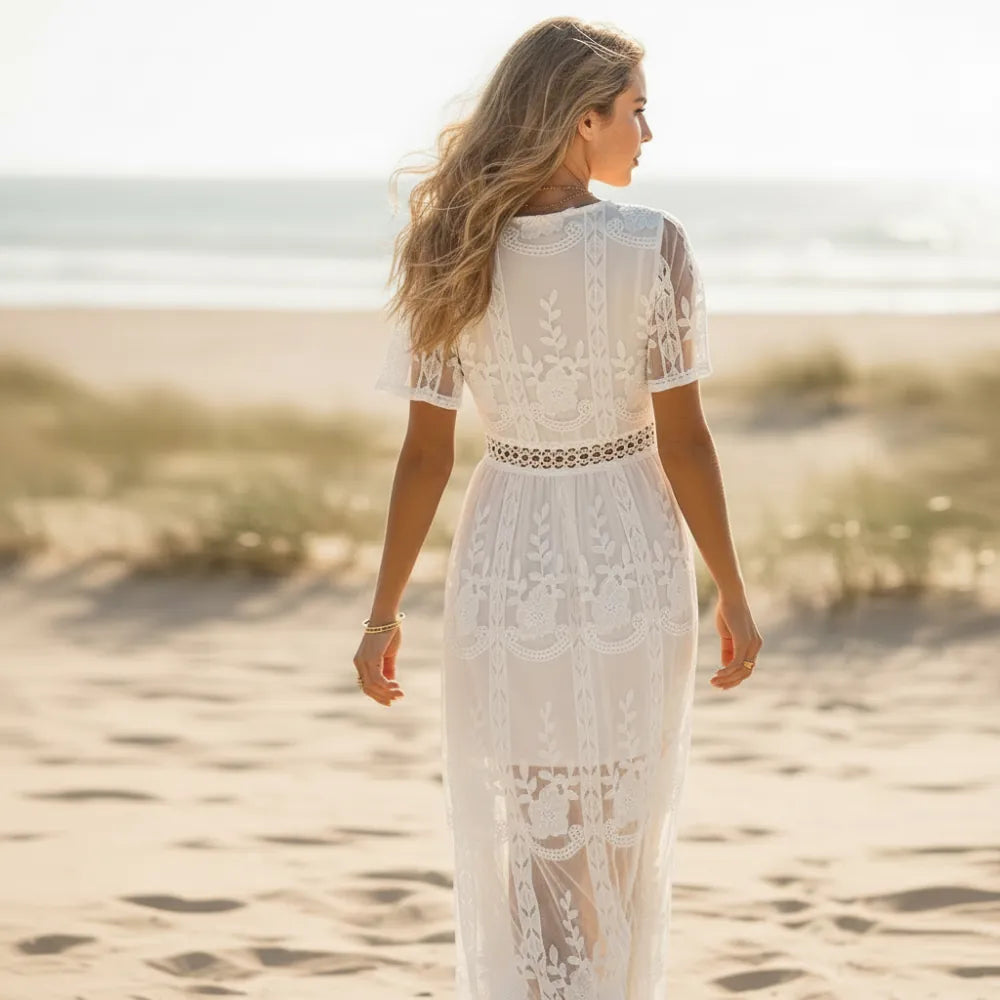 White - A woman standing on a sandy beach at sunset wearing a white lace maxi dress with a plunging V-neckline, short flutter sleeves, and a sheer embroidered skirt.