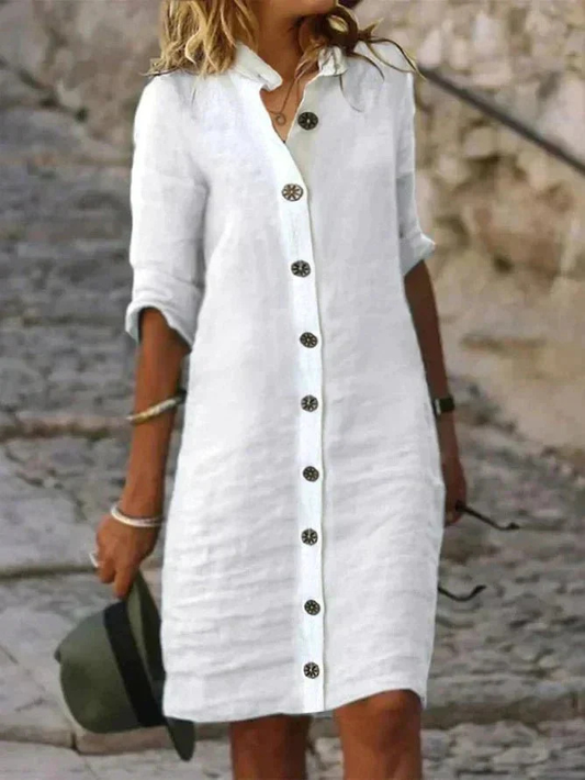 White - Woman walking outdoors wearing a long-sleeve, button-front linen midi dress with a collar, holding a hat.