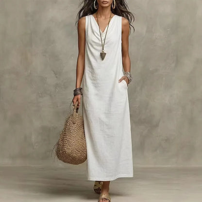 White - A woman wearing a sleeveless, floor-length maxi sundress featuring a draped cowl neckline and side pockets, holding a woven straw tote bag