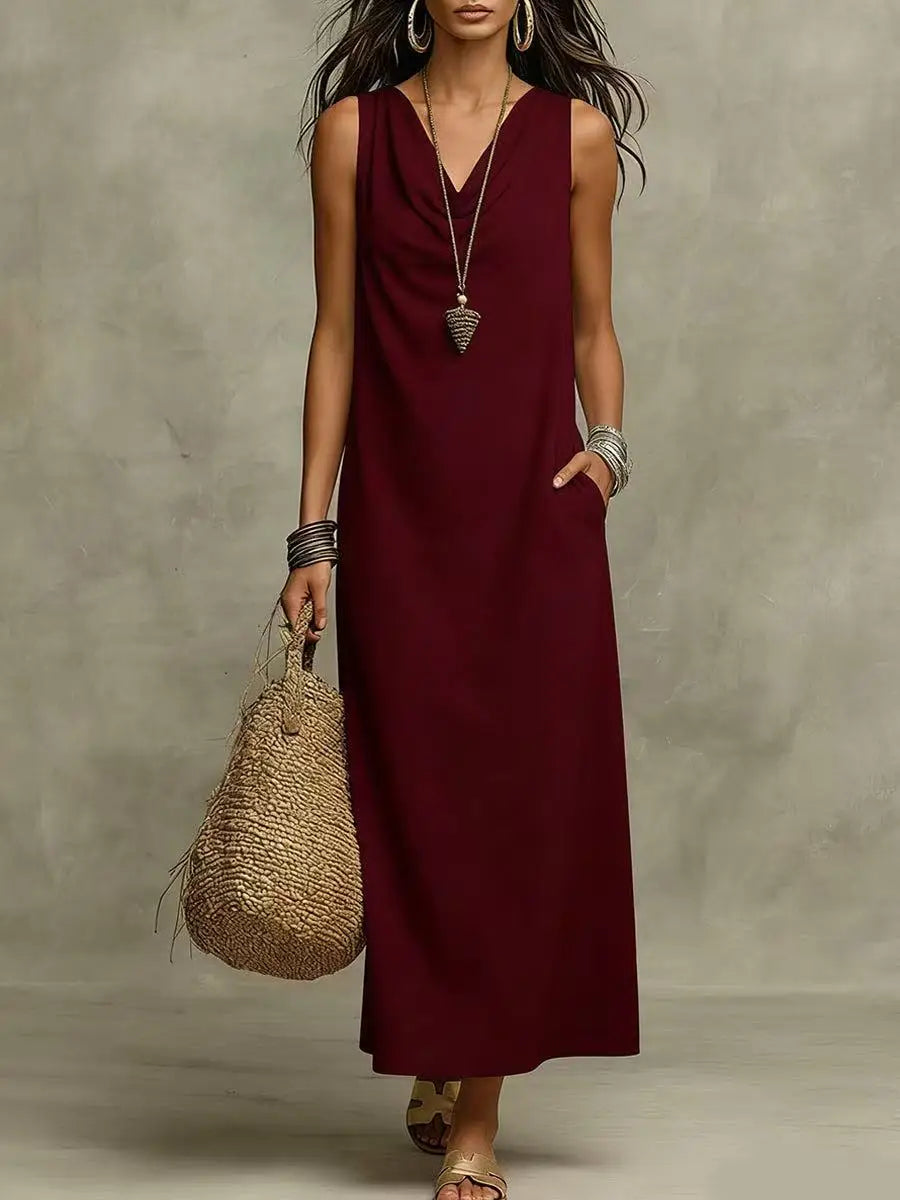 Burgundy - A woman wearing a sleeveless, floor-length maxi sundress featuring a draped cowl neckline and side pockets, holding a woven straw tote bag