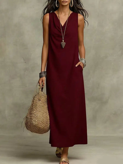 Burgundy - A woman wearing a sleeveless, floor-length maxi sundress featuring a draped cowl neckline and side pockets, holding a woven straw tote bag