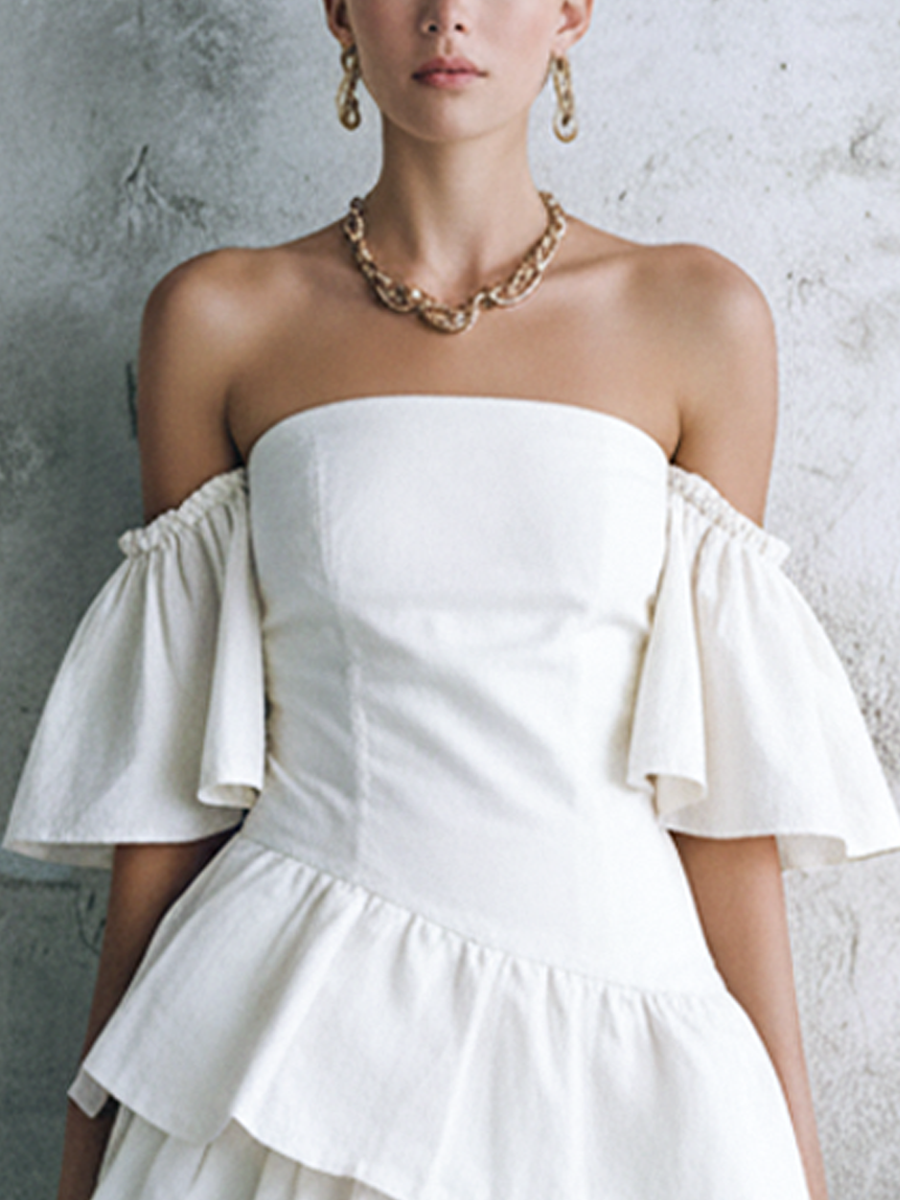 White - Woman wearing a structured off-shoulder tiered mini dress with draped sleeves, holding a small woven bag against a textured stone wall.