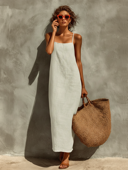 White - Woman wearing a textured, relaxed-fit spaghetti strap maxi dress, holding a large woven tote bag against a concrete wall.