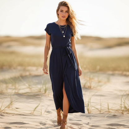 Dark Blue - A woman walks on a sandy beach wearing a black matte jersey maxi dress with cap sleeves, a self-tie belt, and a wrap-front skirt.