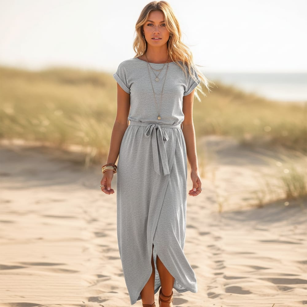 Gray - A woman walks on a sandy beach wearing a black matte jersey maxi dress with cap sleeves, a self-tie belt, and a wrap-front skirt.