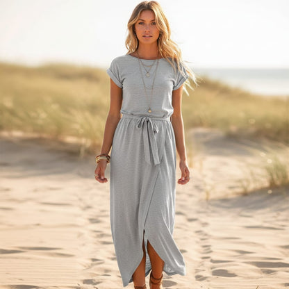 Gray - A woman walks on a sandy beach wearing a black matte jersey maxi dress with cap sleeves, a self-tie belt, and a wrap-front skirt.