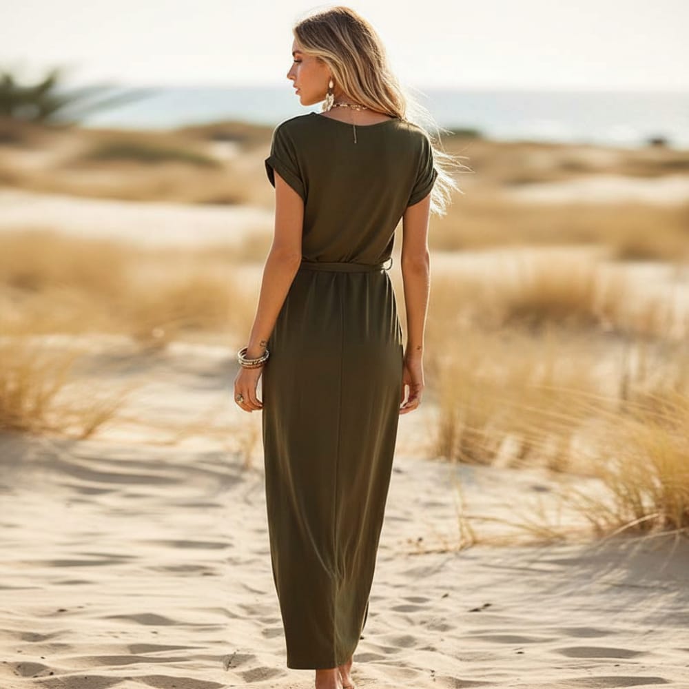Gray - A woman walks on a sandy beach wearing a black matte jersey maxi dress with cap sleeves, a self-tie belt, and a wrap-front skirt.