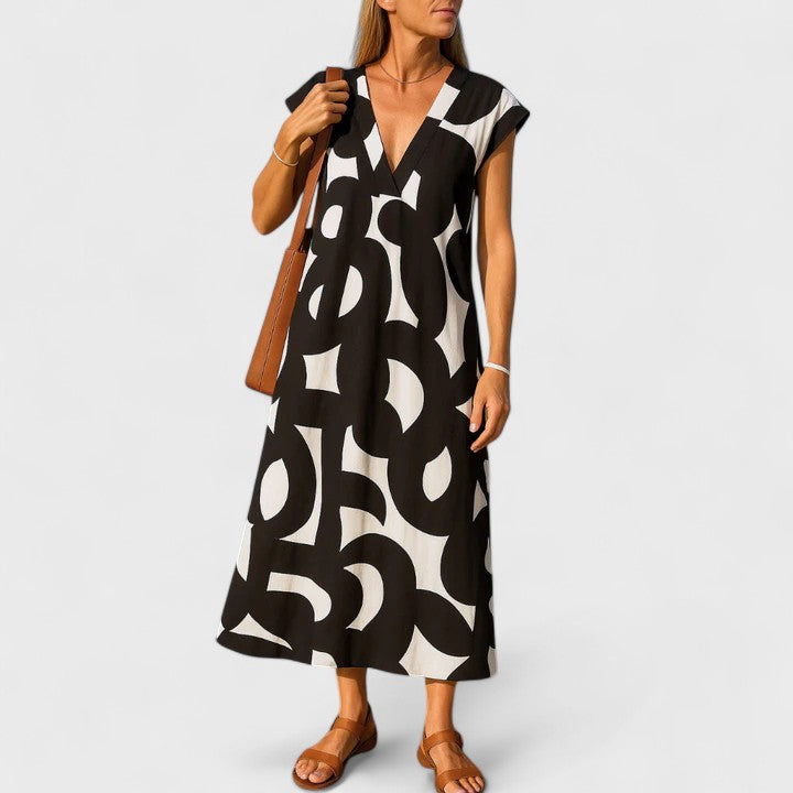 A woman wearing a black and white abstract graphic print midi dress with a V-neckline, cap sleeves, and a relaxed shift silhouette, styled with brown leather sandals and a shoulder bag.