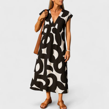A woman wearing a black and white abstract graphic print midi dress with a V-neckline, cap sleeves, and a relaxed shift silhouette, styled with brown leather sandals and a shoulder bag.