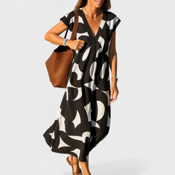 Black - A woman wearing a black and white abstract graphic print midi dress with a V-neckline, cap sleeves, and a relaxed shift silhouette, styled with brown leather sandals and a shoulder bag.