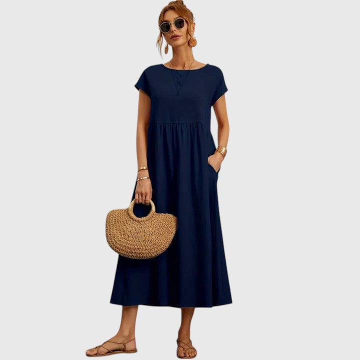 Dark Blue - A woman wearing a navy blue, cap-sleeve midi sundress with an empire waist and side pockets, styled with a woven straw handbag and flat sandals.