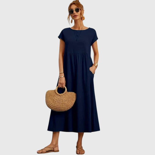 Dark Blue - A woman wearing a navy blue, cap-sleeve midi sundress with an empire waist and side pockets, styled with a woven straw handbag and flat sandals.