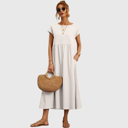 White - A woman wearing a navy blue, cap-sleeve midi sundress with an empire waist and side pockets, styled with a woven straw handbag and flat sandals.