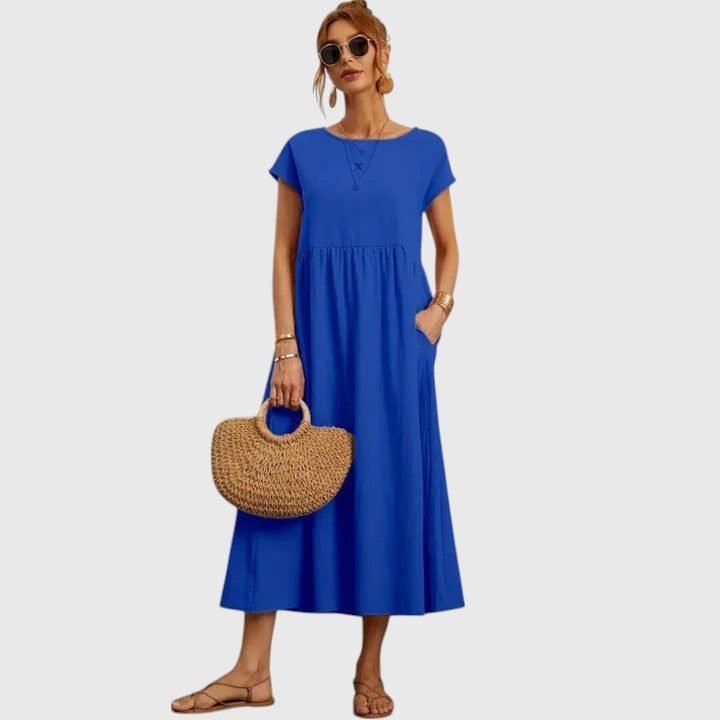 Blue - A woman wearing a navy blue, cap-sleeve midi sundress with an empire waist and side pockets, styled with a woven straw handbag and flat sandals.