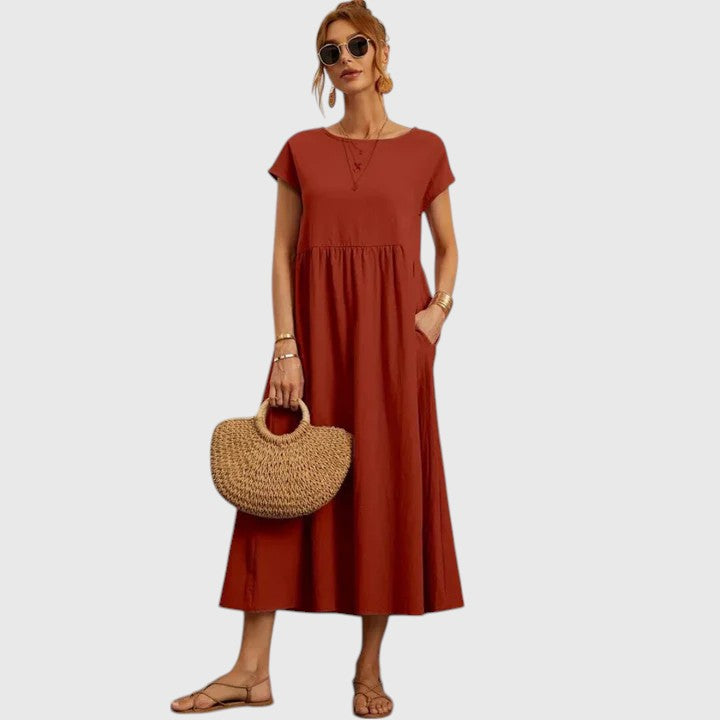 Rusty - A woman wearing a navy blue, cap-sleeve midi sundress with an empire waist and side pockets, styled with a woven straw handbag and flat sandals.