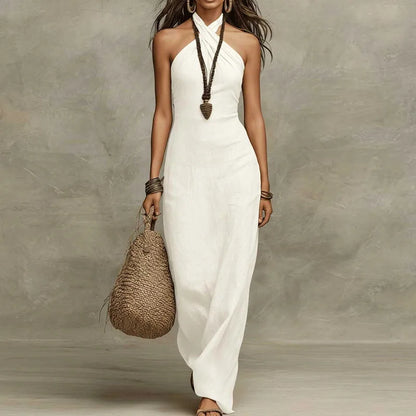 White - A woman wearing a floor-length white linen-blend maxi dress with a criss-cross halter neckline, styled with a woven straw bag and minimalist accessories against a textured gray background.