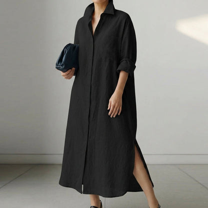 Black - A woman wearing a black, relaxed-fit linen maxi shirt dress with a collared neckline, button-down front, rolled-up sleeves, and high side slits, paired with a dark blue clutch bag.