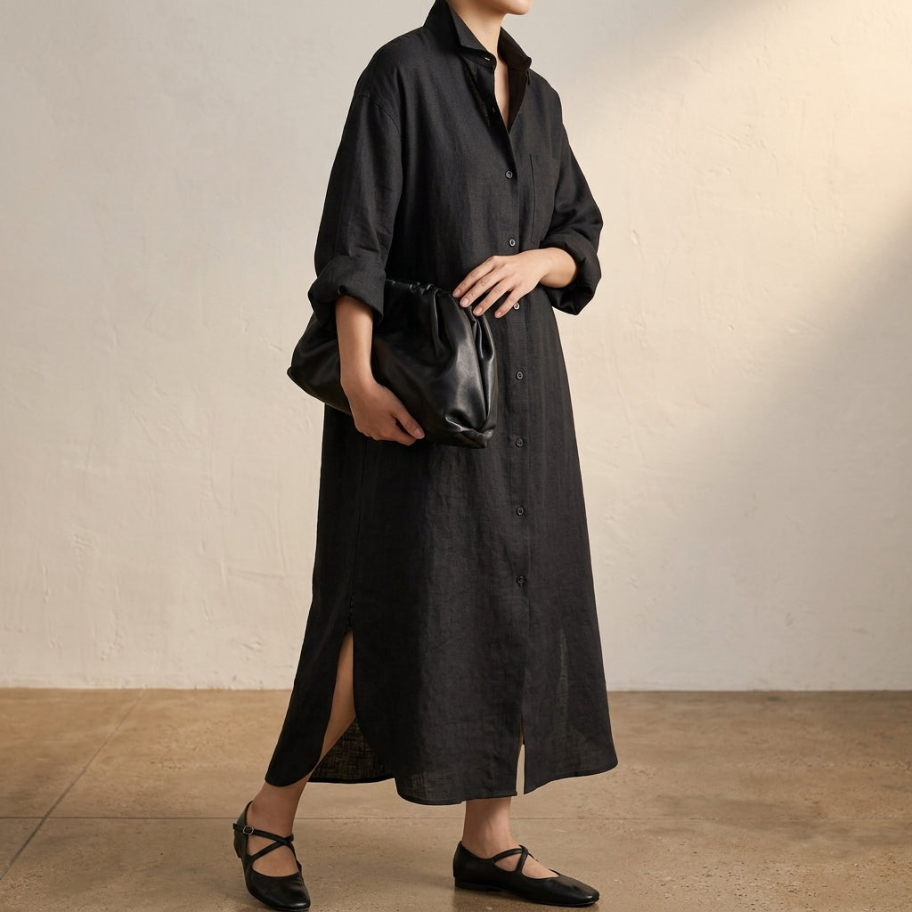 Beige - A woman wearing a black, relaxed-fit linen maxi shirt dress with a collared neckline, button-down front, rolled-up sleeves, and high side slits, paired with a dark blue clutch bag.