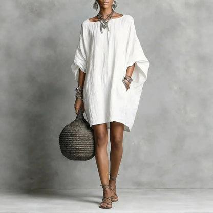White - A woman wearing an oversized white crinkled linen mini sundress with dramatic dolman sleeves, a boat neckline, and side pockets, paired with strappy sandals.