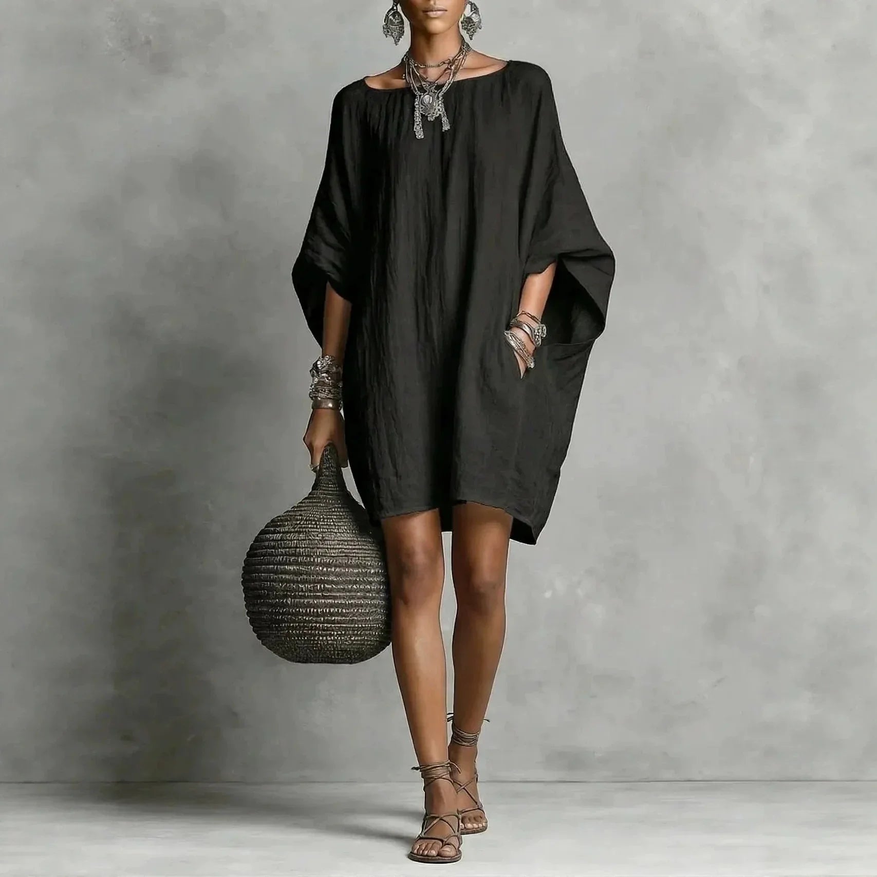 Black - A woman wearing an oversized white crinkled linen mini sundress with dramatic dolman sleeves, a boat neckline, and side pockets, paired with strappy sandals.