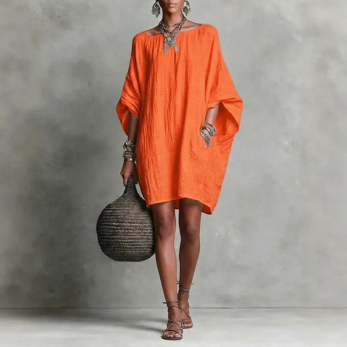Orange - A woman wearing an oversized white crinkled linen mini sundress with dramatic dolman sleeves, a boat neckline, and side pockets, paired with strappy sandals.