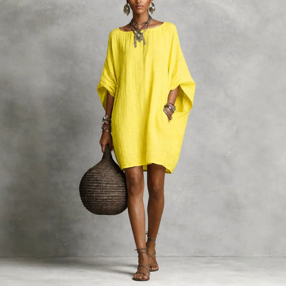 Yellow - A woman wearing an oversized white crinkled linen mini sundress with dramatic dolman sleeves, a boat neckline, and side pockets, paired with strappy sandals.