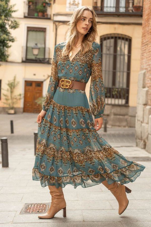 3XL - A woman wearing a teal and brown paisley print chiffon maxi dress with long bishop sleeves, a V-neckline, and a tiered ruffle skirt, styled with a brown leather belt and tan knee-high boots.
