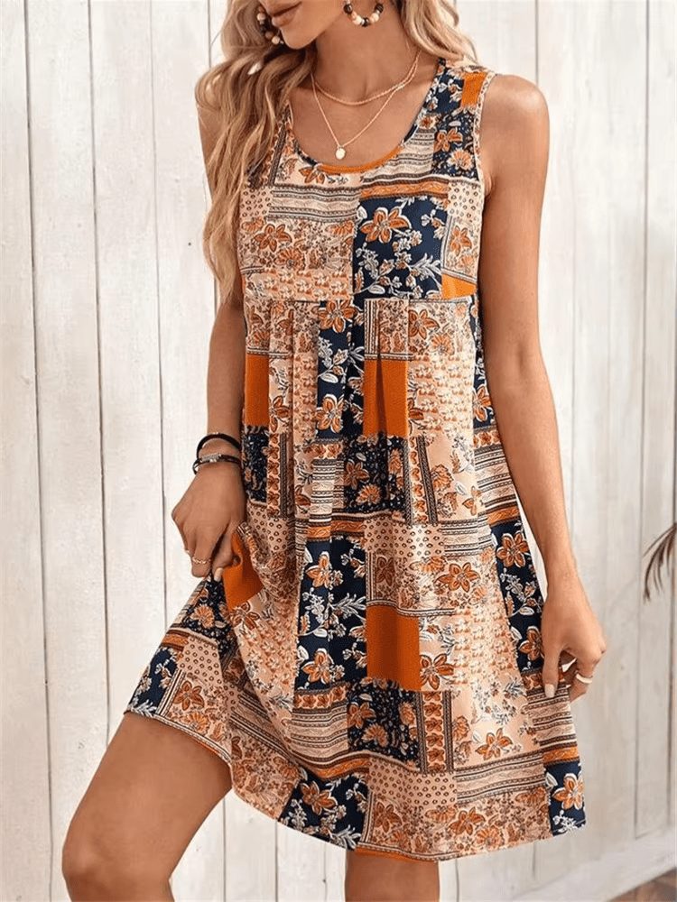 A woman wearing a sleeveless mini dress featuring a multi-colored patchwork floral print in orange, navy, and cream, styled with a scoop neckline and a gathered empire waist.