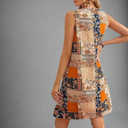 Orange - A woman wearing a sleeveless mini dress featuring a multi-colored patchwork floral print in orange, navy, and cream, styled with a scoop neckline and a gathered empire waist.