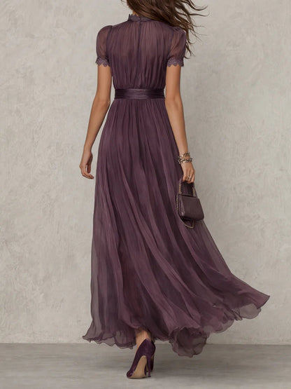 Purple - A woman wearing a flowing, deep mauve pleated chiffon maxi dress with a high keyhole neckline, lace-trimmed puff sleeves, and a banded waist.