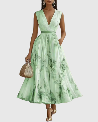 Green - Model wearing a sleeveless, plunging V-neck midi dress in pleated off-white chiffon with a blue and gold watercolor floral print, featuring a cinched blue waist band and a flowy A-line skirt with pockets.