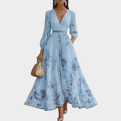 White - Model wearing a sleeveless, plunging V-neck midi dress in pleated off-white chiffon with a blue and gold watercolor floral print, featuring a cinched blue waist band and a flowy A-line skirt with pockets.