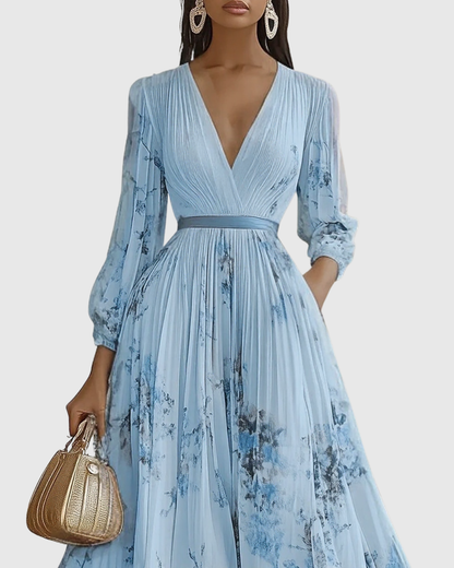 Blue - Model wearing a sleeveless, plunging V-neck midi dress in pleated off-white chiffon with a blue and gold watercolor floral print, featuring a cinched blue waist band and a flowy A-line skirt with pockets.
