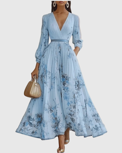 Blue - Model wearing a sleeveless, plunging V-neck midi dress in pleated off-white chiffon with a blue and gold watercolor floral print, featuring a cinched blue waist band and a flowy A-line skirt with pockets.