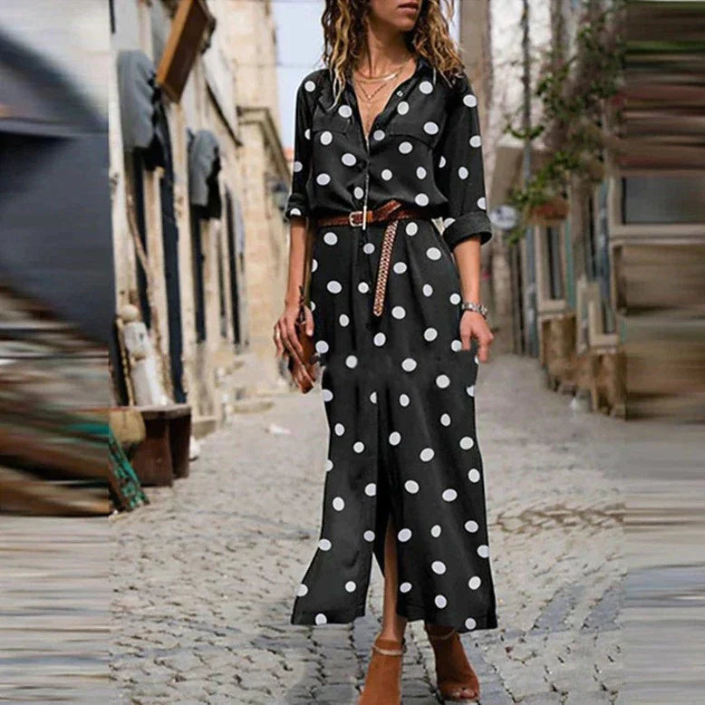 Black - Woman wearing an olive green maxi shirt dress with white polka dots, featuring a collared button-down front, rolled-up long sleeves, and a brown belt, walking on a cobblestone street.