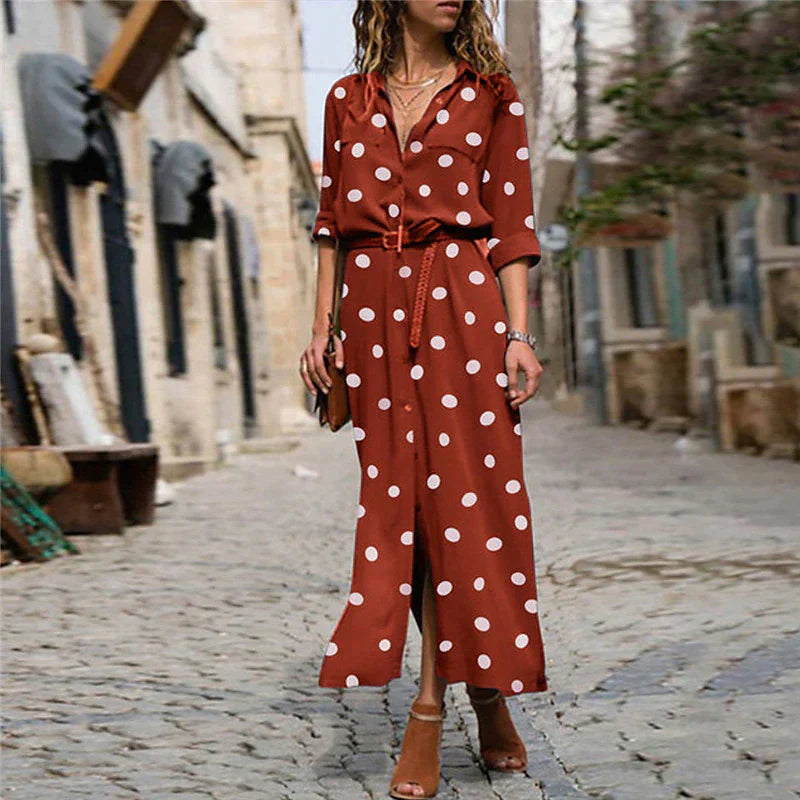 Red - Woman wearing an olive green maxi shirt dress with white polka dots, featuring a collared button-down front, rolled-up long sleeves, and a brown belt, walking on a cobblestone street.
