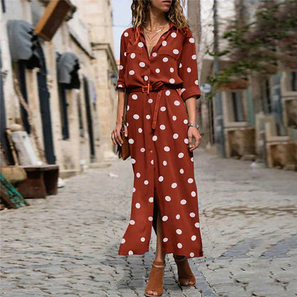 Red - Woman wearing an olive green maxi shirt dress with white polka dots, featuring a collared button-down front, rolled-up long sleeves, and a brown belt, walking on a cobblestone street.