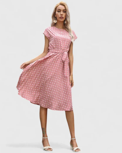 Pink - A woman wearing a dusty pink midi dress with white polka dots, featuring cap sleeves, a belted waist, and a pleated skirt.