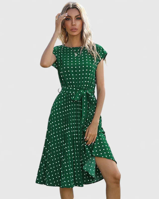 Green - A woman wearing a dusty pink midi dress with white polka dots, featuring cap sleeves, a belted waist, and a pleated skirt.