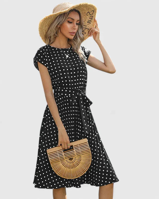 Black - A woman wearing a dusty pink midi dress with white polka dots, featuring cap sleeves, a belted waist, and a pleated skirt.