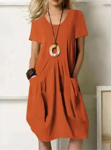 Rust - Woman wearing a solid color short sleeve knee-length shift dress featuring front pleats and two large patch pockets