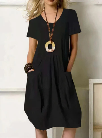 Black - Woman wearing a solid color short sleeve knee-length shift dress featuring front pleats and two large patch pockets