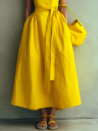 Yellow - Woman wearing a square-neck midi dress with wide straps, a self-tie waist, and a voluminous A-line skirt, holding a matching handbag against a textured grey background.