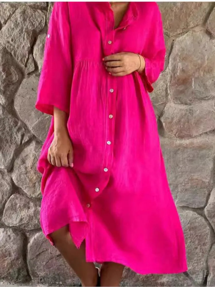 Pink - A woman wearing a vibrant fuchsia pink linen midi dress with a button-down front, roll-tab sleeves, and an empire waist, standing against a stone wall.