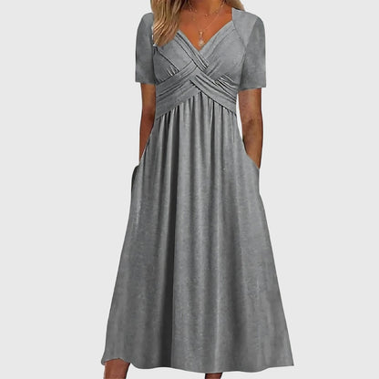 Gray - A woman wearing a cornflower blue midi-length jersey dress featuring a criss-cross ruched bodice, short sleeves, and a flowy skirt with side pockets.