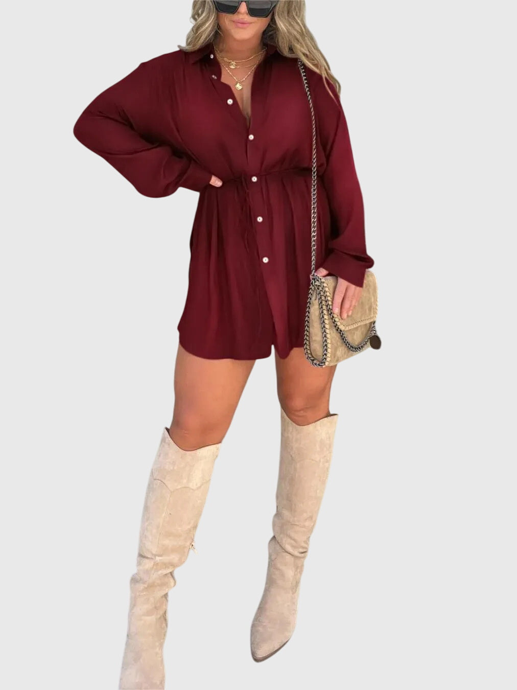 Wine Red - A woman wearing a long-sleeve burgundy satin romper with a button-down front, pointed collar, and cinched waist, styled with knee-high beige boots.