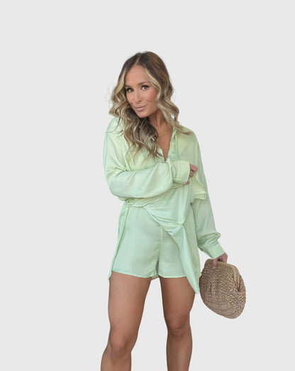 Melon - A woman wearing a long-sleeve burgundy satin romper with a button-down front, pointed collar, and cinched waist, styled with knee-high beige boots.