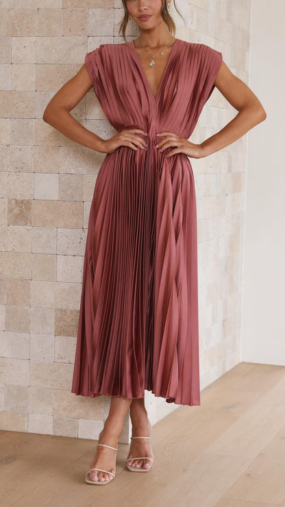 A woman wearing a dusty rose pleated satin midi dress with a deep V-neckline, cap sleeves, and a cinched waist, standing against a textured stone wall.