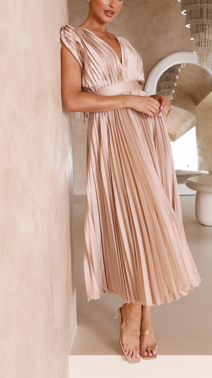 Champagne - A woman wearing a dusty rose pleated satin midi dress with a deep V-neckline, cap sleeves, and a cinched waist, standing against a textured stone wall.